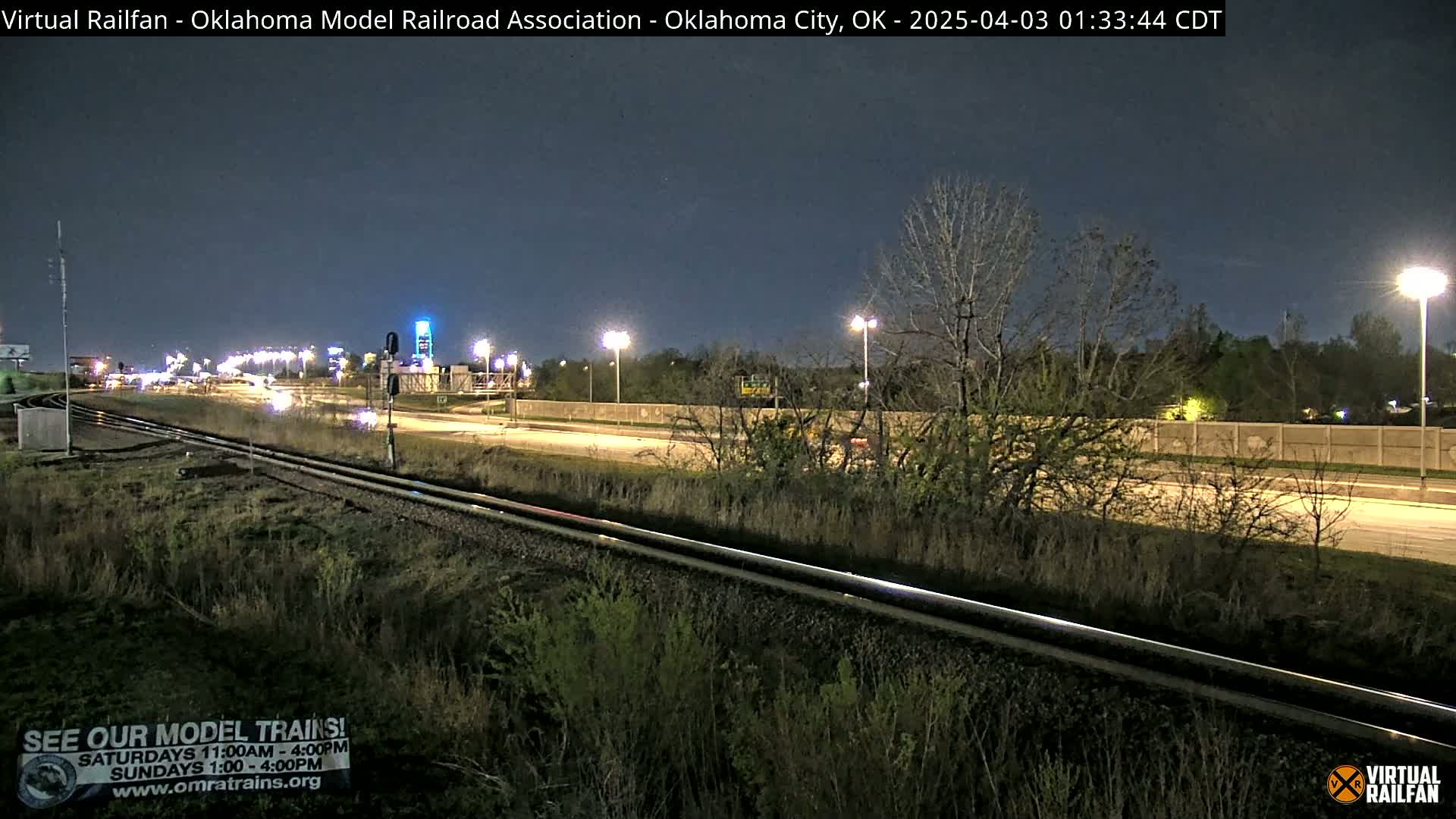 A nighttime view of a highway and railroad tracks, with sparse vegetation and streetlights illuminating the scene.
