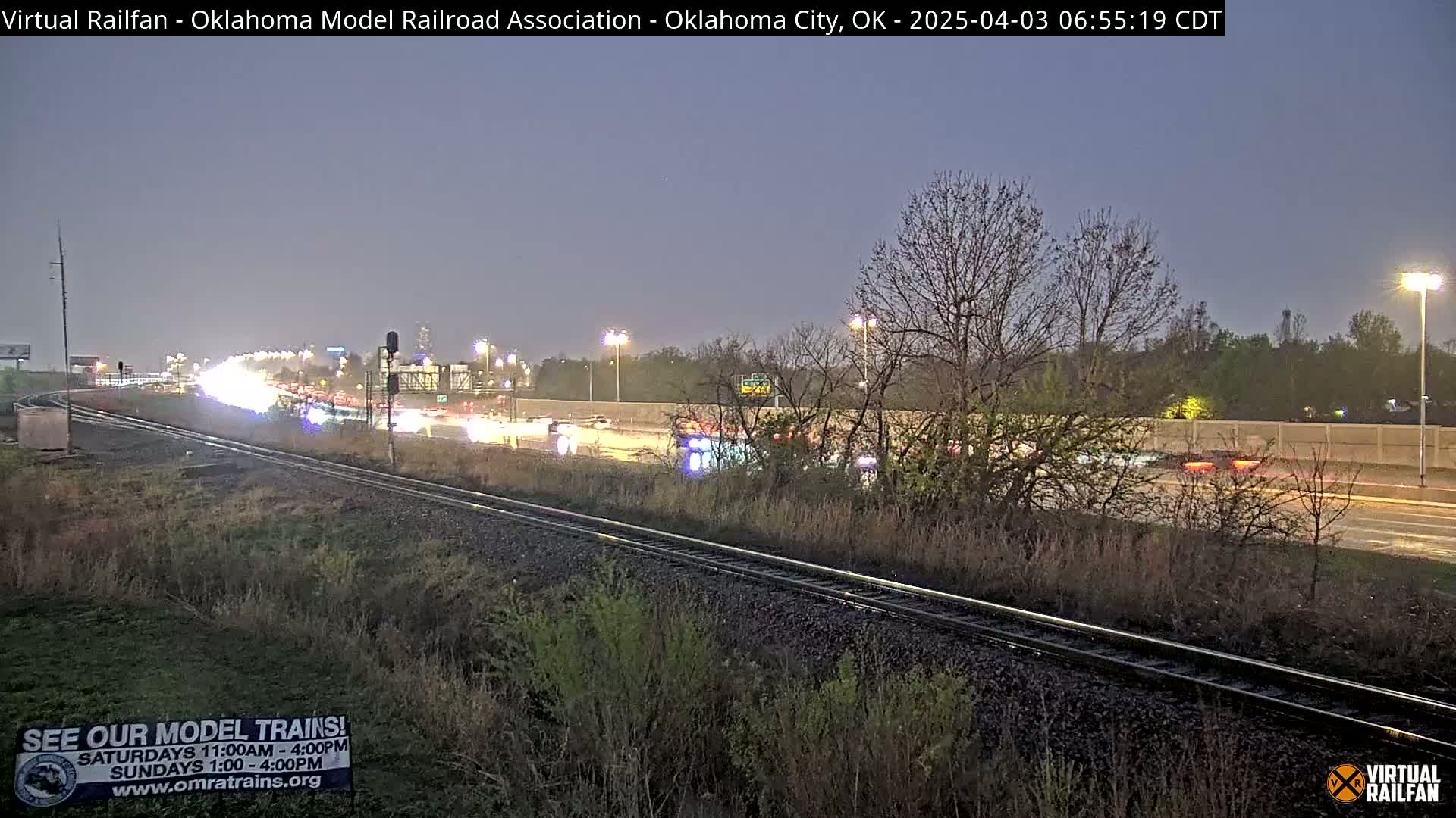 A nighttime view of a highway with moderate traffic beside a set of train tracks, under a dusky sky.