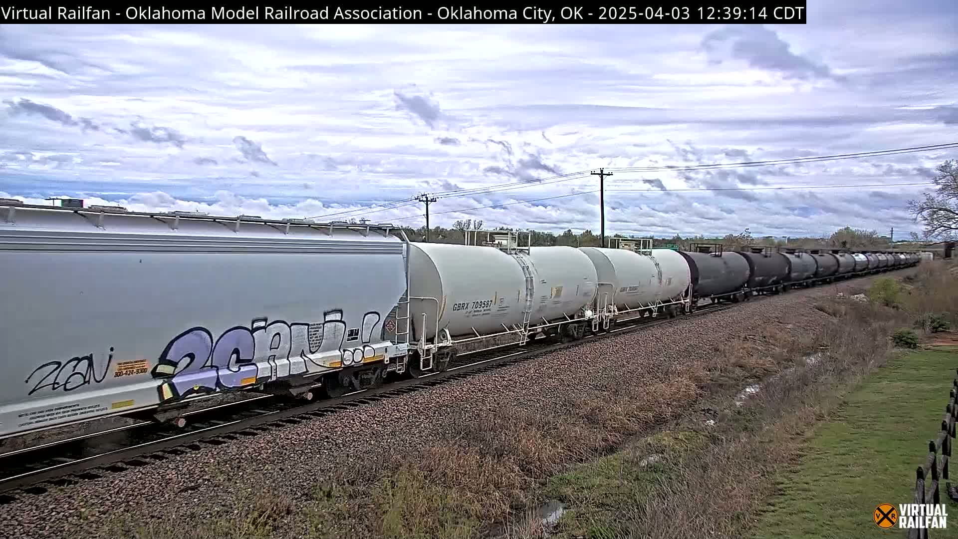 A long freight train with light gray and dark gray tanker cars passes by under a partly cloudy sky.