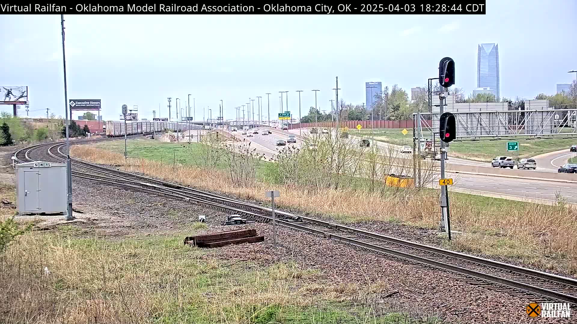 A long freight train is visible on a railway line next to a highway with light traffic under a partly cloudy sky.