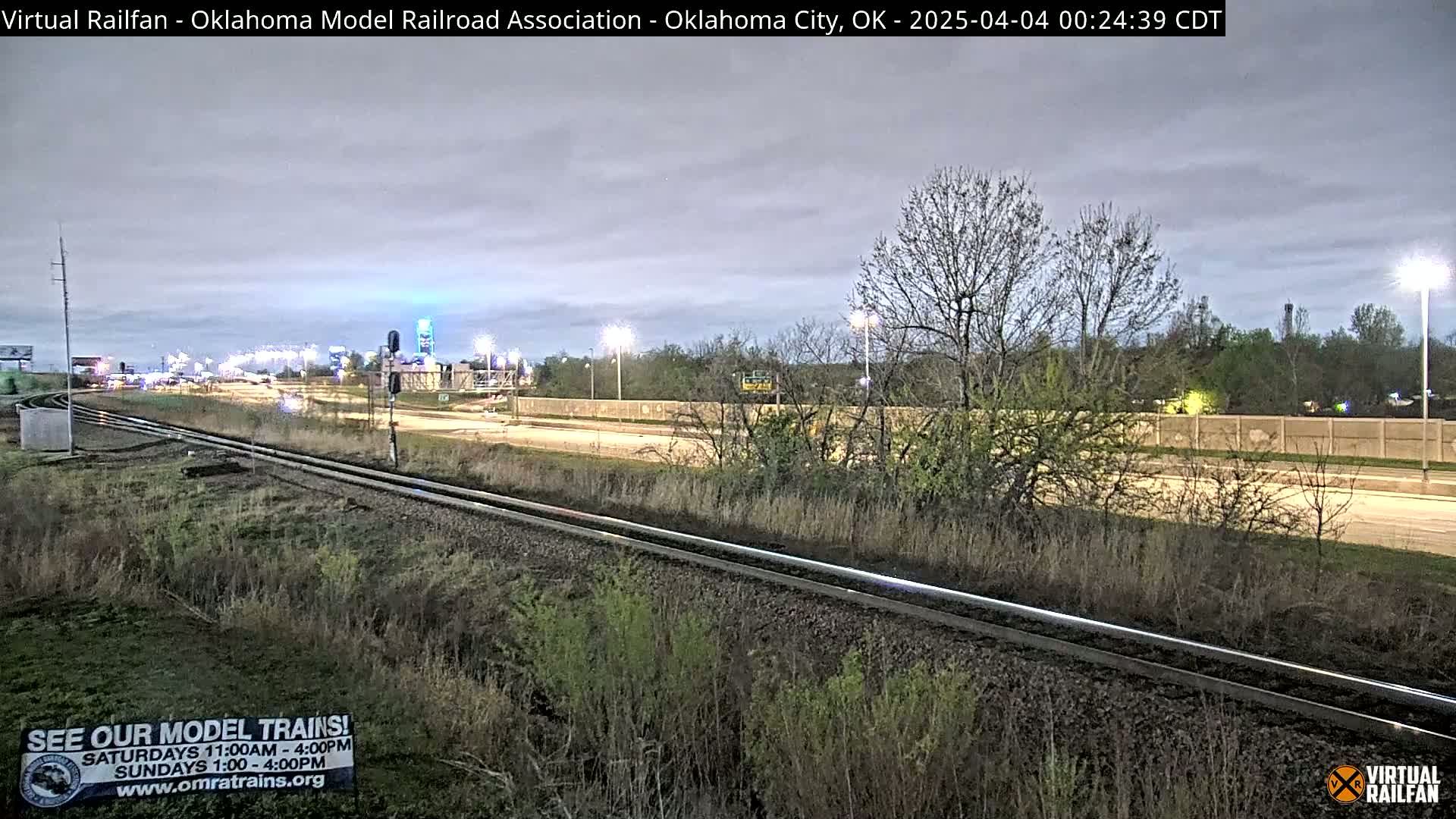 A nighttime view of a highway and parallel train tracks, with sparse, leafless trees and undergrowth alongside, under a cloudy sky.