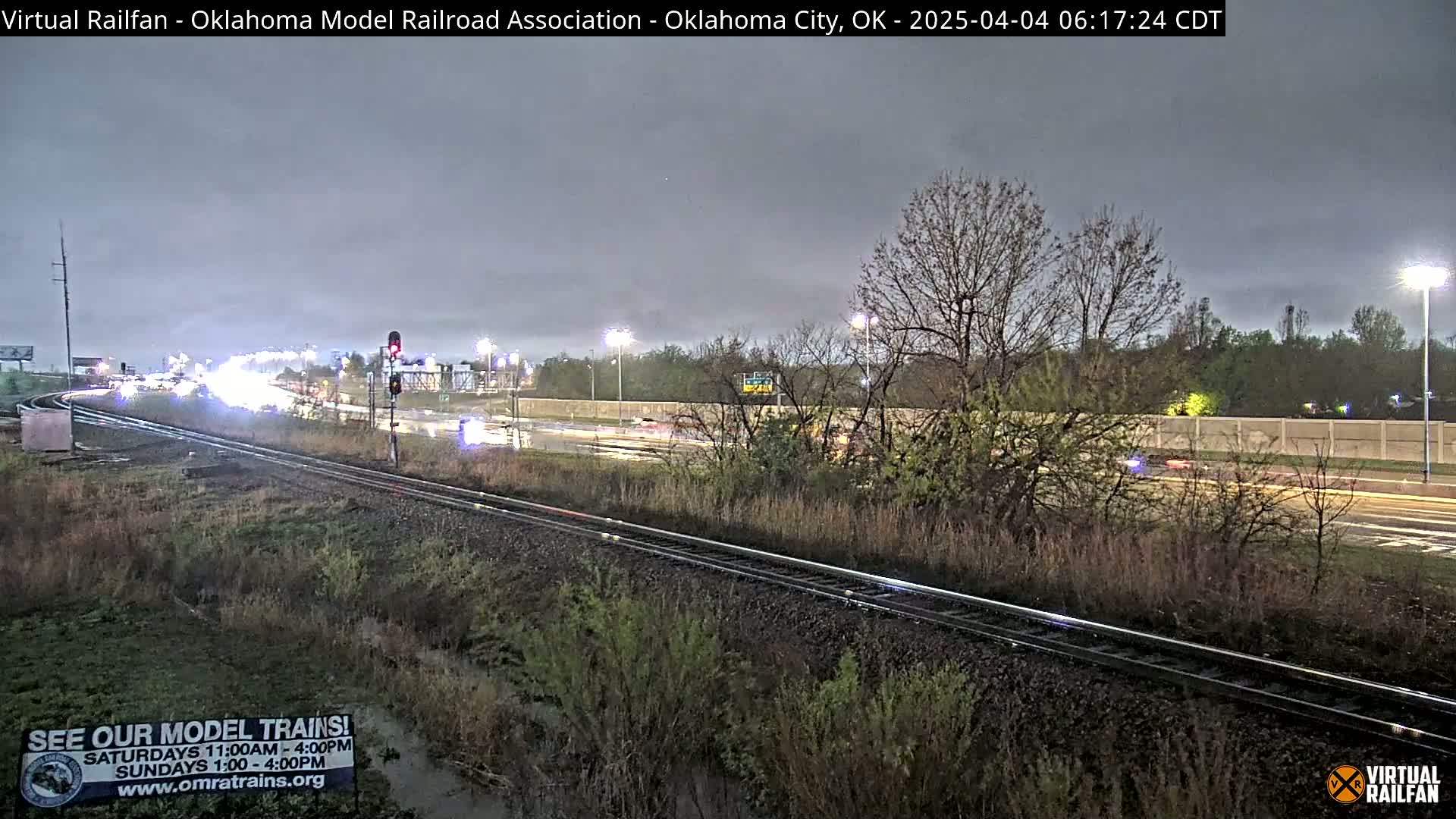 A wet, overcast scene shows a railroad track next to a highway with cars driving on it at dusk.