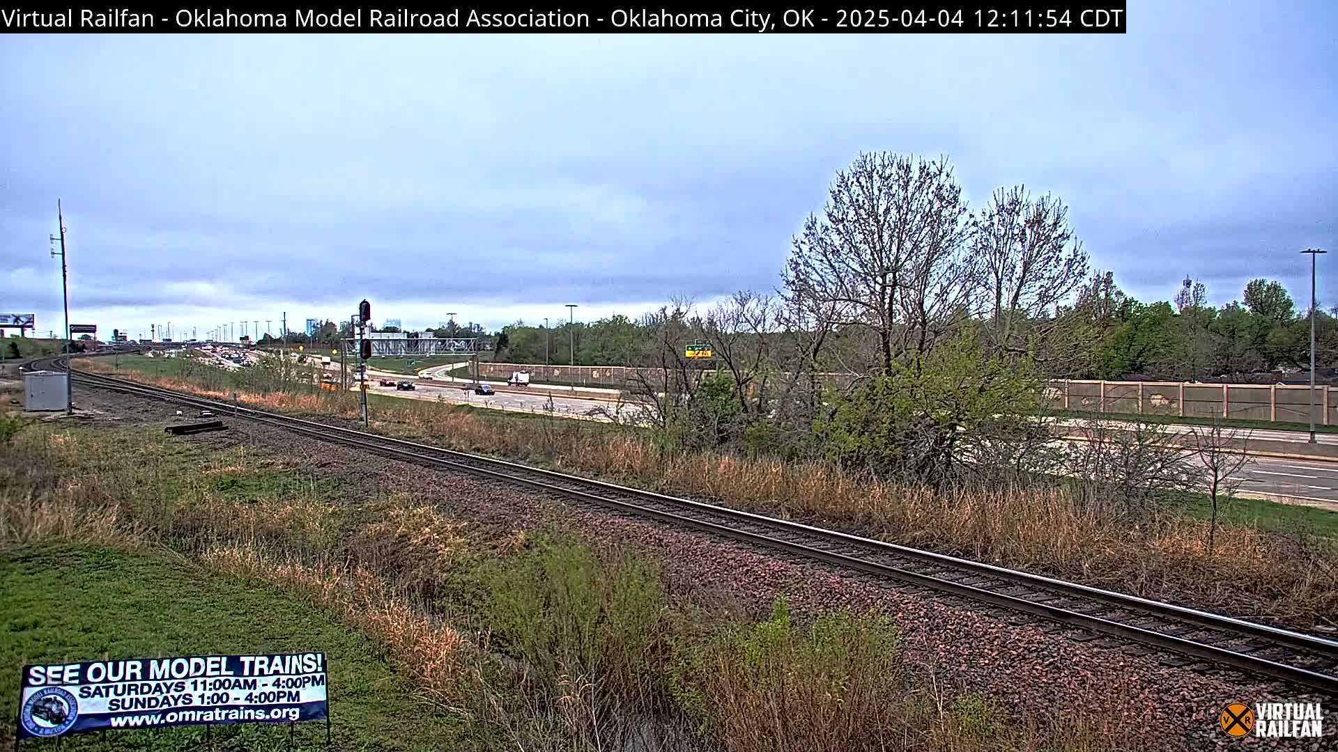 A mostly sunny day shows a railroad track running alongside a highway with moderate traffic, bordered by vegetation including bare trees and grass.
