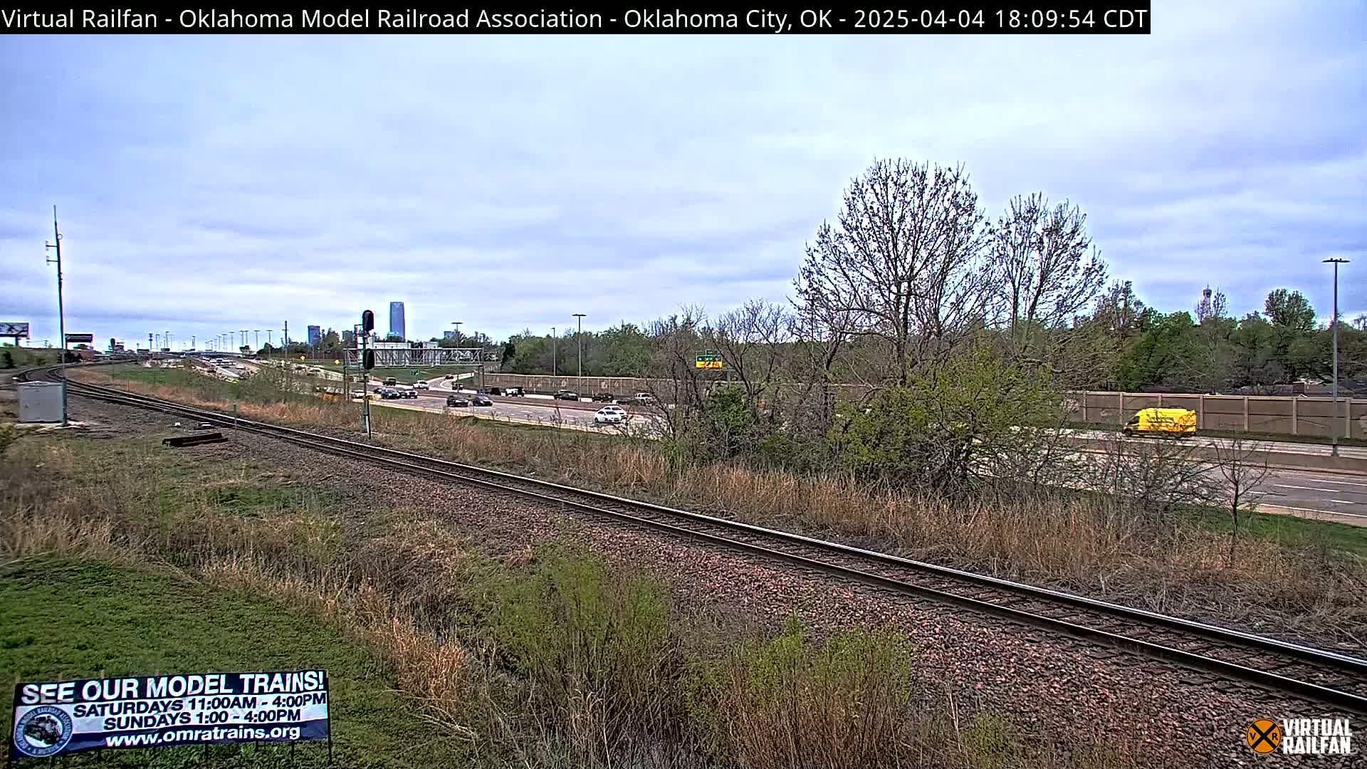 A mostly clear sky overlooks railroad tracks adjacent to a highway with moderate traffic, featuring sparse, budding trees and scrubby vegetation.