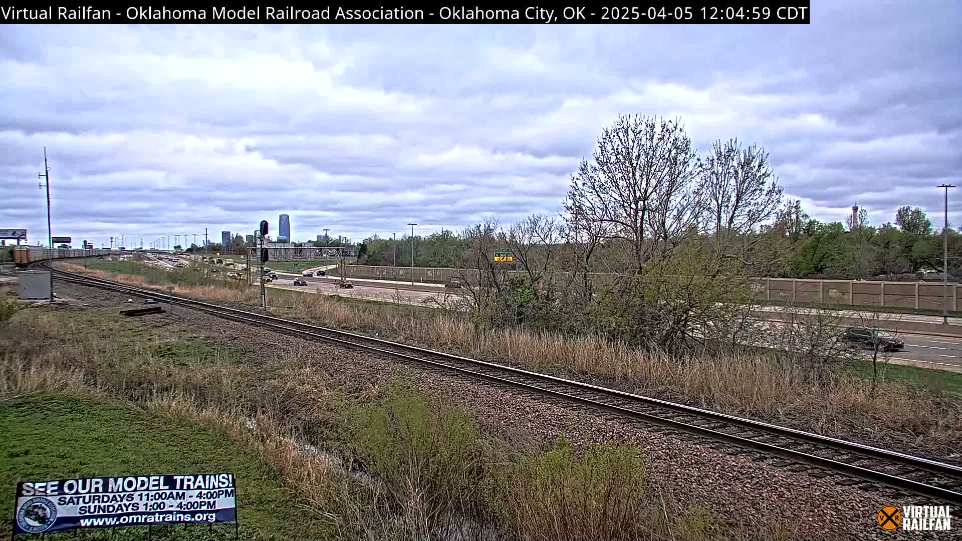 A mostly cloudy day reveals railroad tracks beside a highway with sparse traffic,  trees budding with early spring leaves, and a distant cityscape.