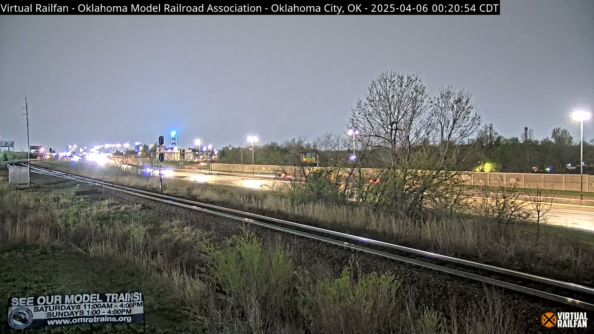 A nighttime view of railroad tracks beside a highway with sparse traffic under a twilight sky.