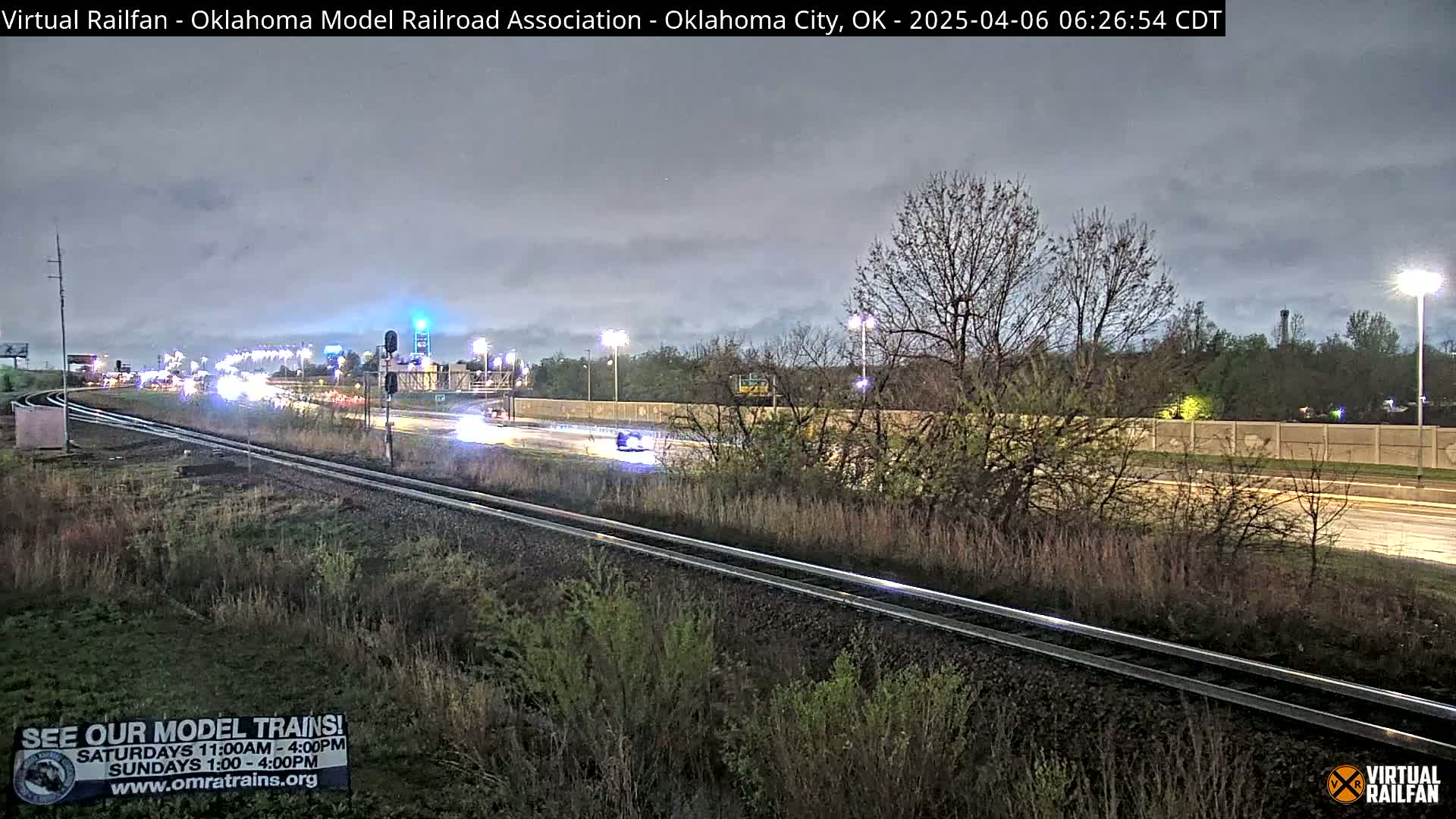 Under a cloudy twilight sky, a highway with moderate traffic runs parallel to a set of railroad tracks beside some vegetation.