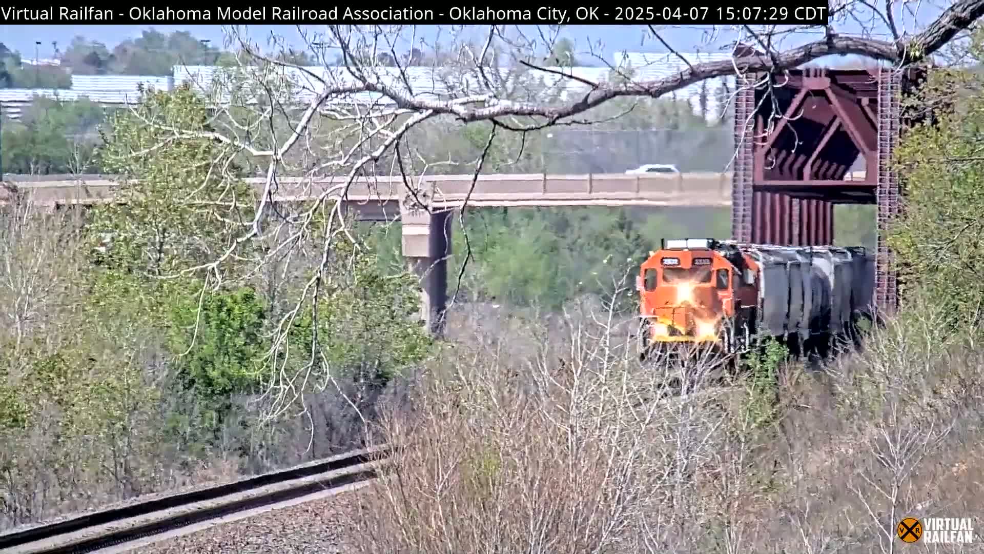 An orange train engine pulling a long freight train passes under a bridge on a sunny day, partially obscured by trees and brush.