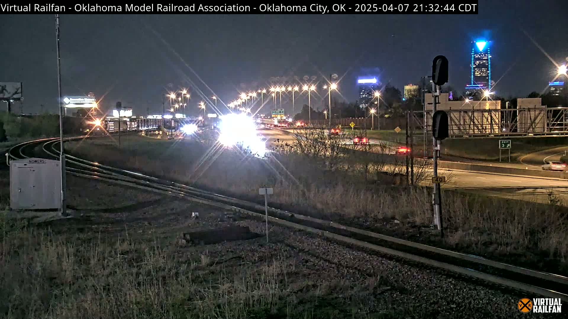 A nighttime view of train tracks curving alongside a highway with cars driving under a bridge and illuminated buildings in the distance.