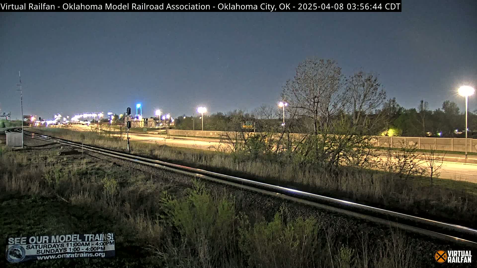 Under a dark night sky, railroad tracks run alongside a highway, both bordered by vegetation and illuminated by streetlights.