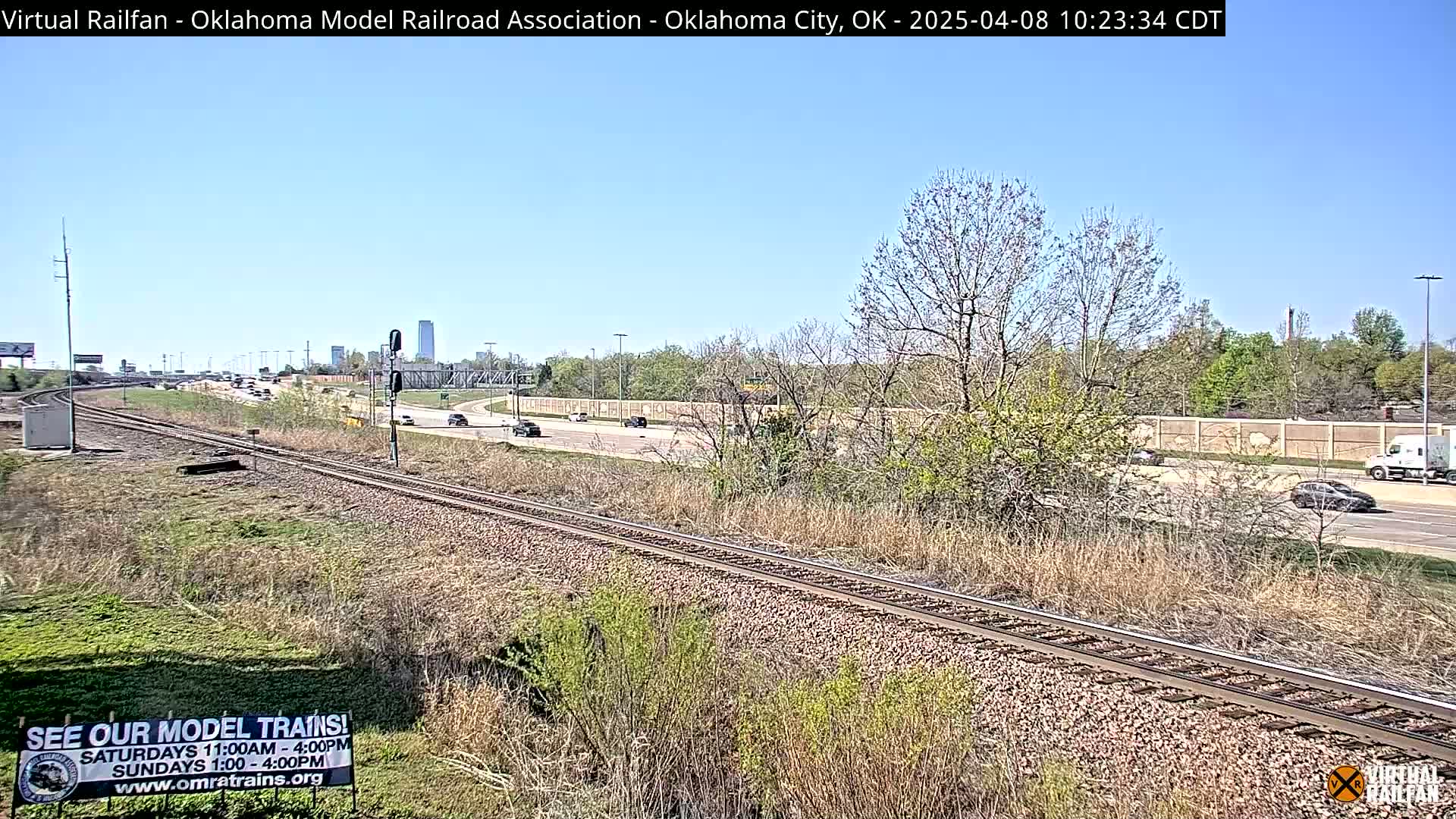 A train track runs parallel to a highway under a clear blue sky, with sparse, early spring vegetation bordering both.
