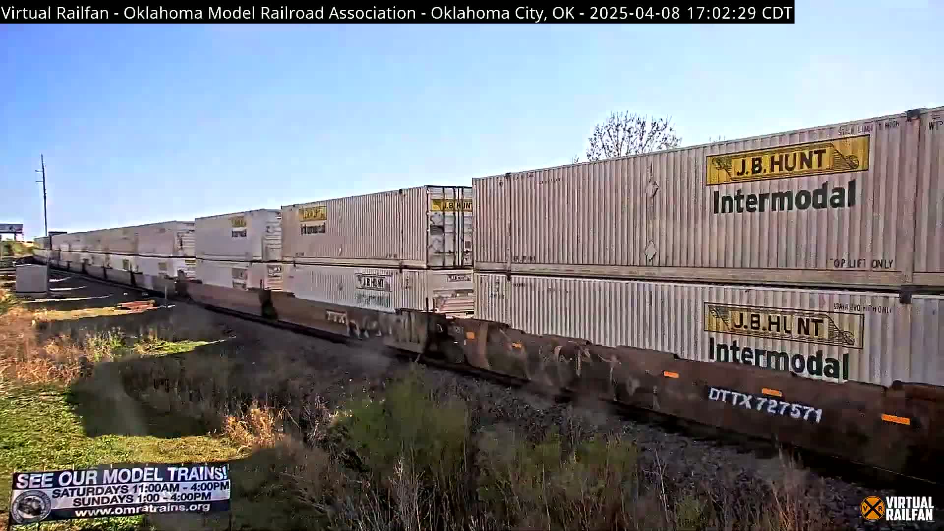 Under a clear blue sky, a long freight train carrying numerous stacked shipping containers moves along a railway track beside dry, sparse vegetation.
