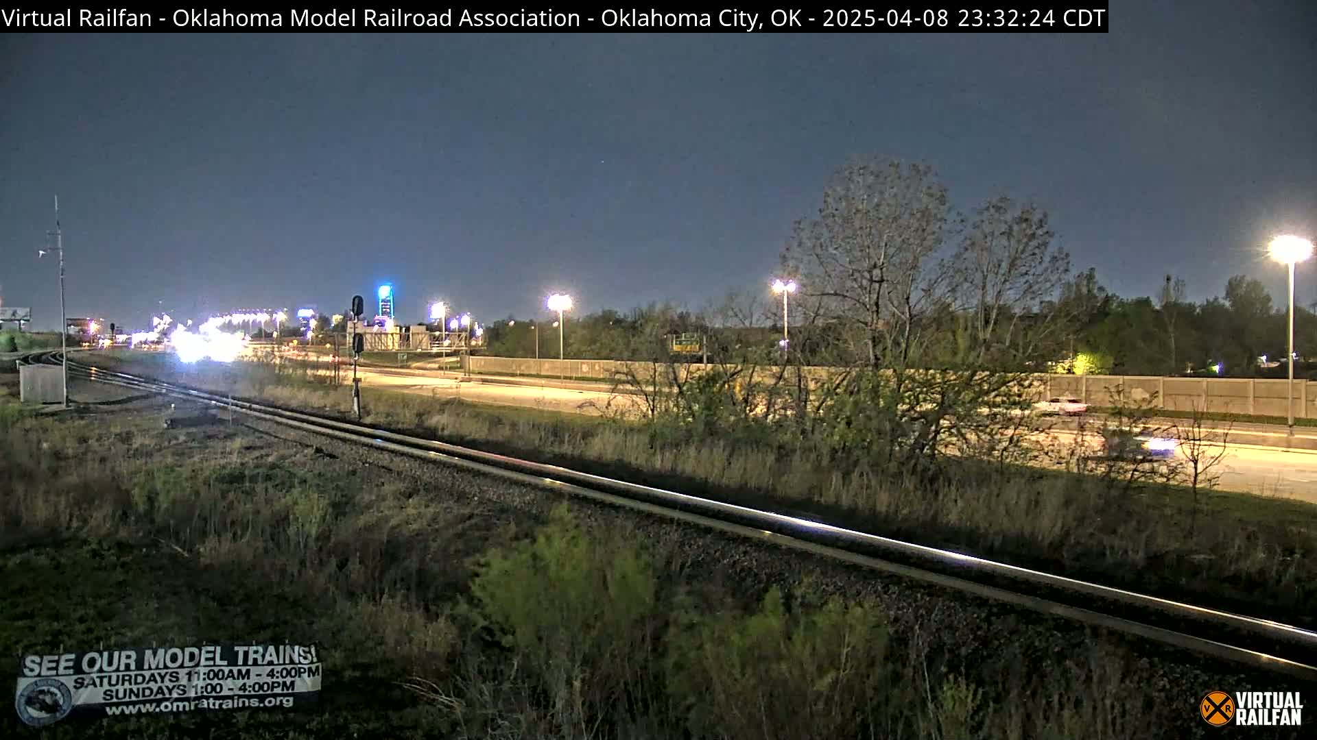 A nighttime view of a highway and parallel train tracks, with sparsely lit vegetation and a few vehicles visible on the road.