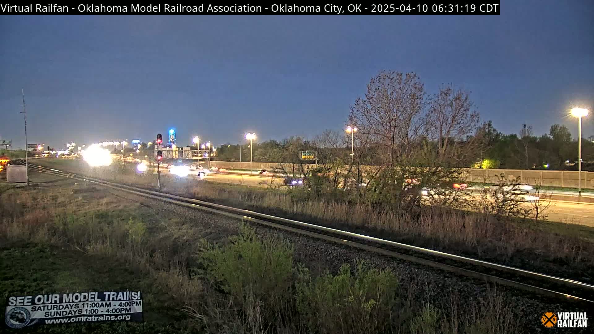 A nighttime view of a highway with light traffic, beside railroad tracks, under a twilight sky.