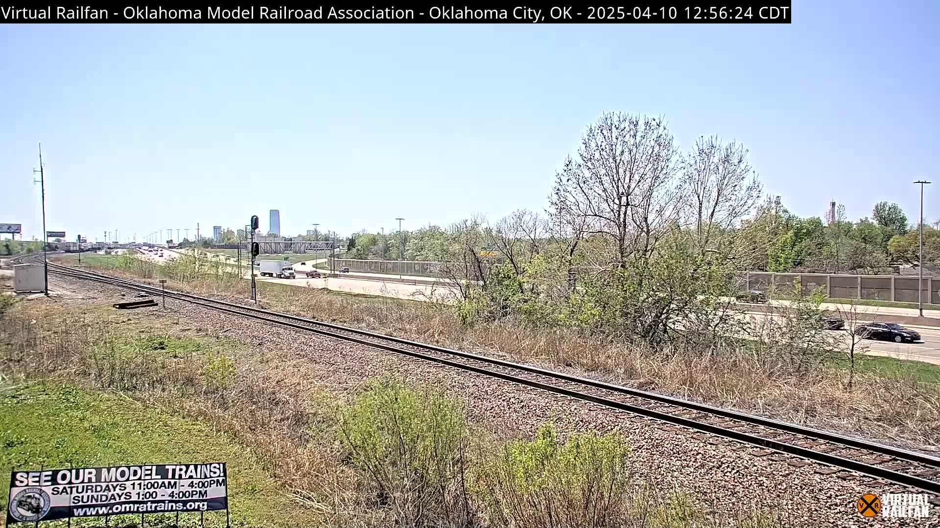 A train track runs parallel to a highway, both bordered by scrubby vegetation under a clear blue sky.