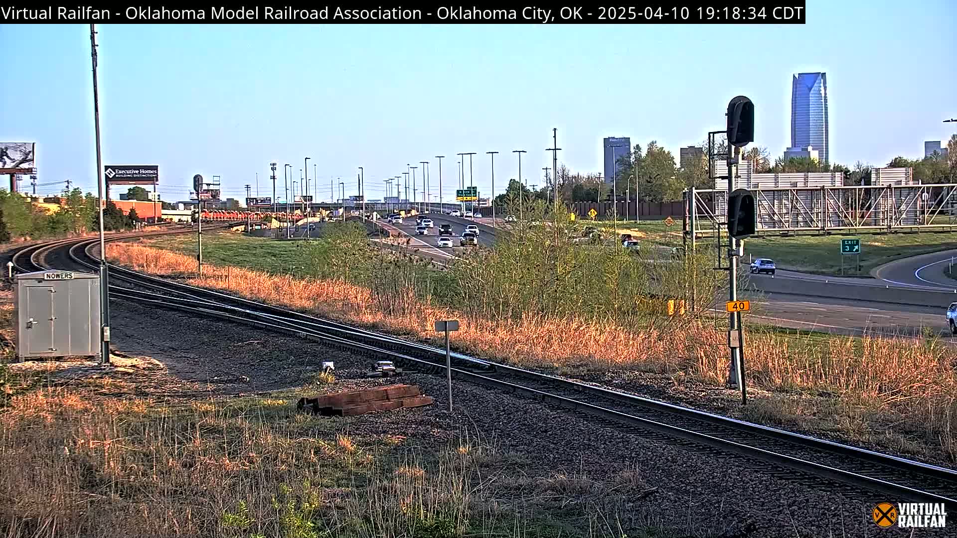 Railroad tracks curve alongside a highway with moderate traffic under a clear daytime sky, and a cityscape is visible in the background.