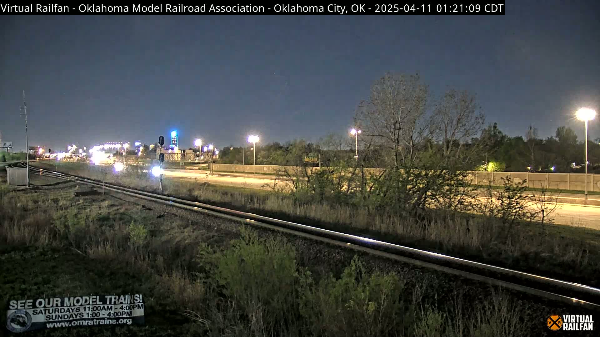 Under a dark night sky, railroad tracks run alongside a highway with sparse traffic, bordered by vegetation and illuminated by streetlights.