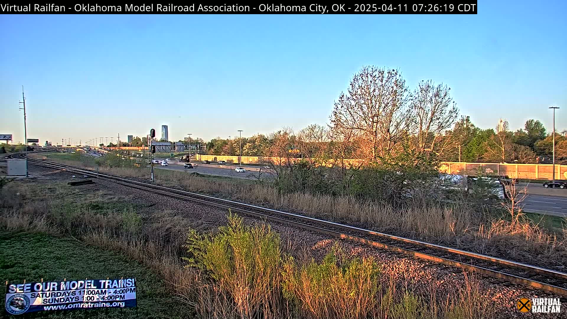 Under a clear blue sky, a railroad track runs parallel to a highway, with cars driving on the road and trees and brush bordering the tracks.