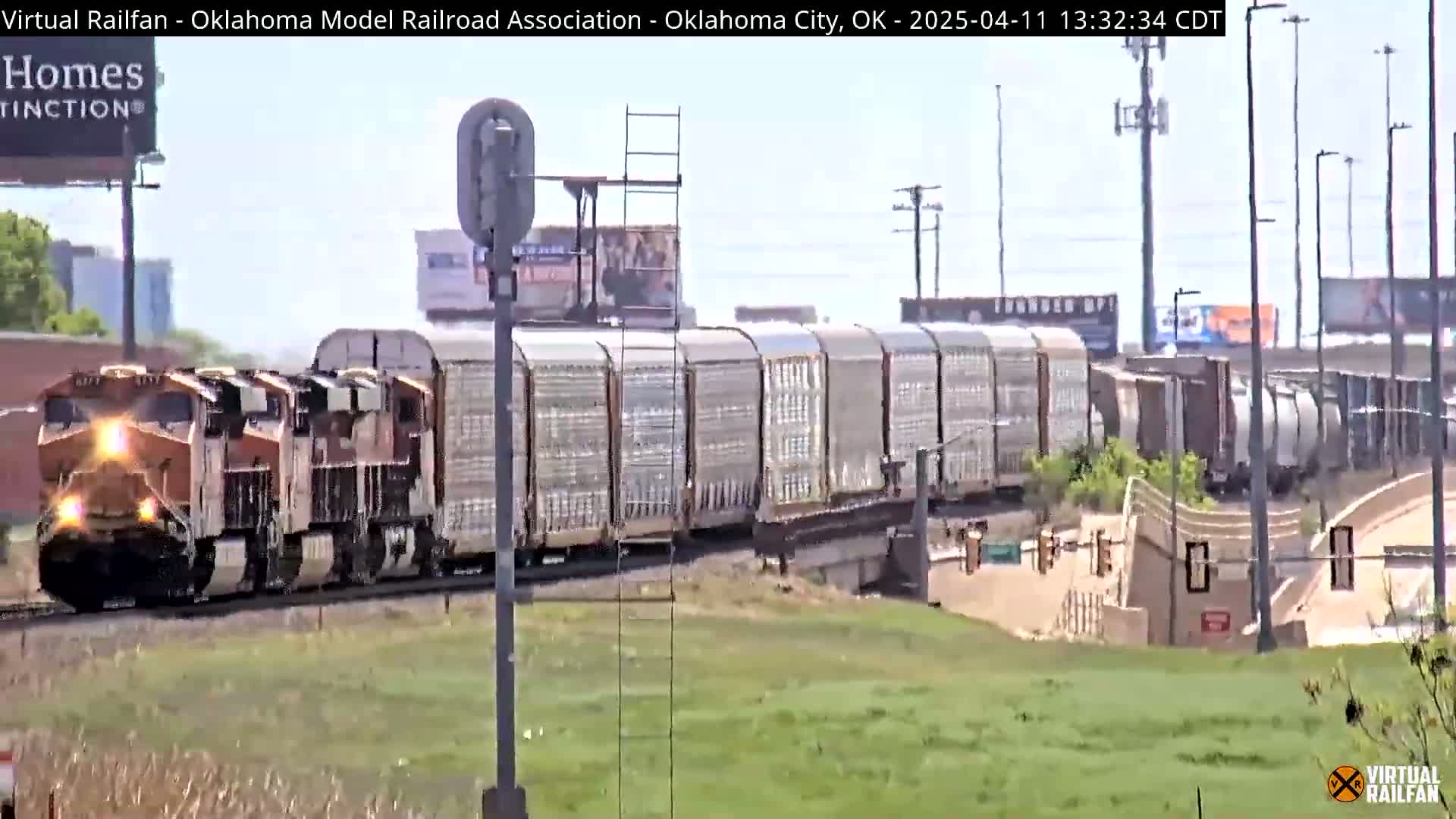 A long freight train with two orange locomotives passes by under a mostly sunny sky.