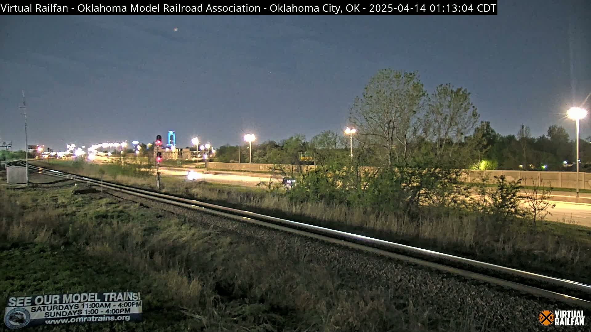 A nighttime view of railroad tracks beside a highway, with sparse vegetation and streetlights illuminating the scene.