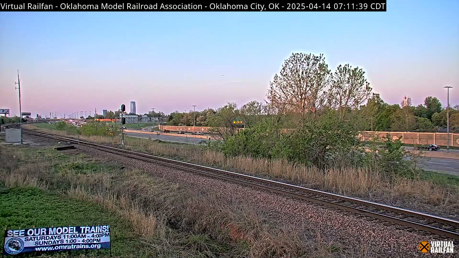 A section of railroad tracks runs alongside a highway in the early evening, with sparse vegetation and trees lining the roadside.