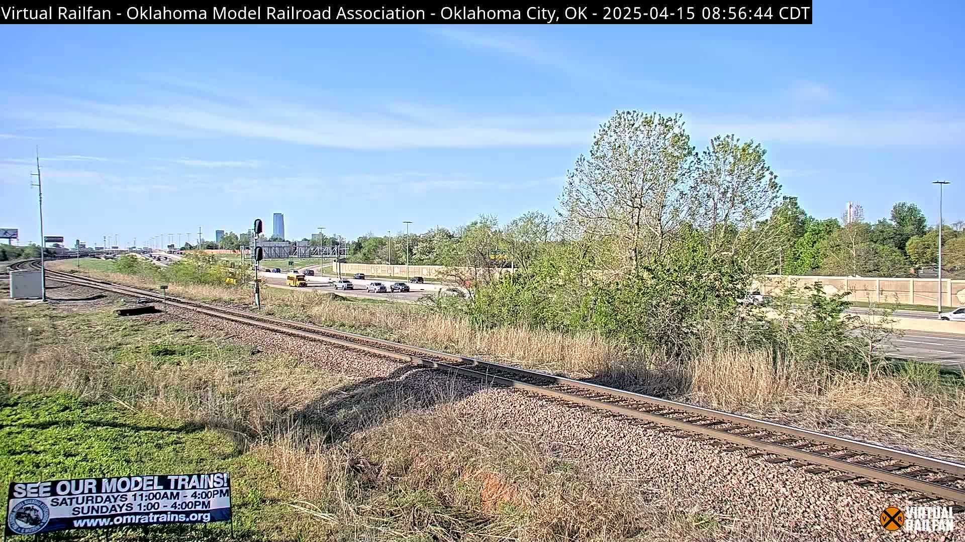 A sunny day shows a railway track next to a highway with cars driving on it, some low-lying vegetation, and trees in the background.