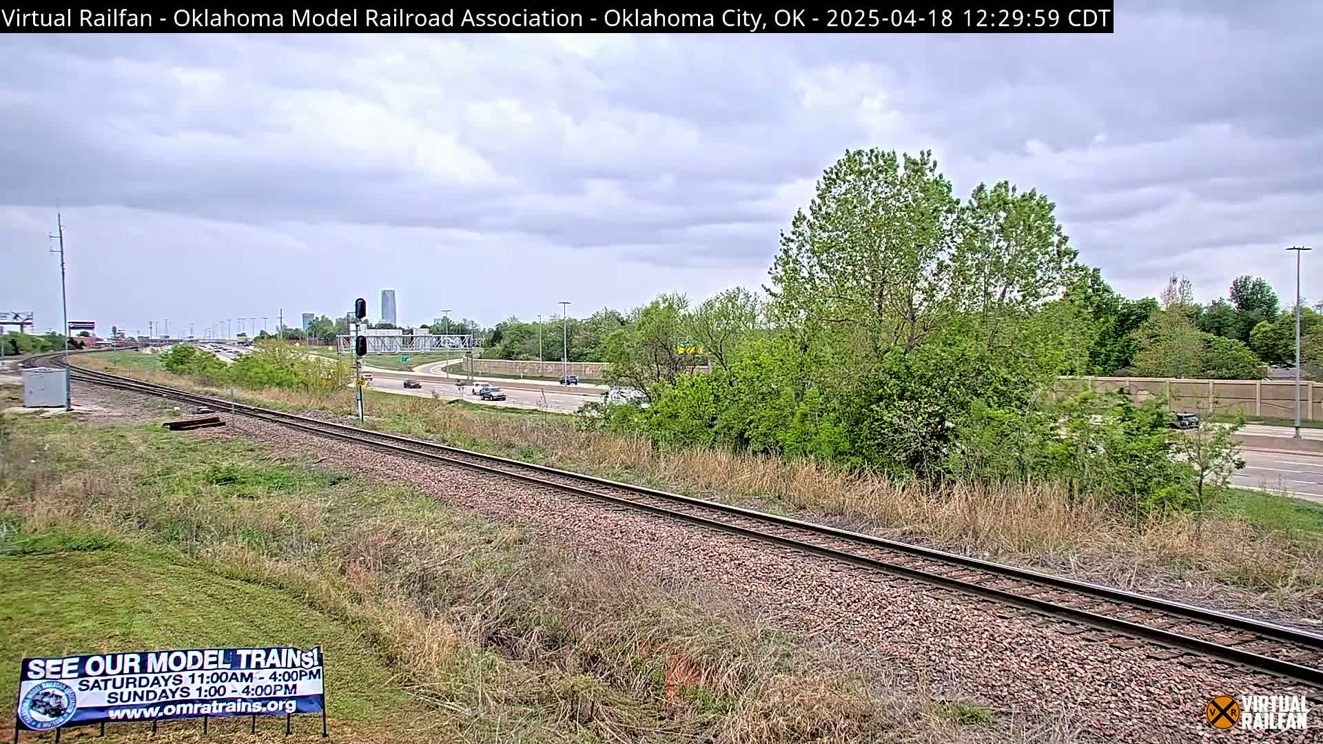 Railroad tracks run alongside a highway with light traffic under a partly cloudy sky, bordered by green vegetation.