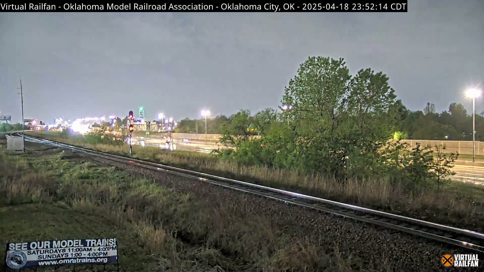 A nighttime view of railroad tracks beside a highway under a dark, overcast sky, with some light rain visible on the road.
