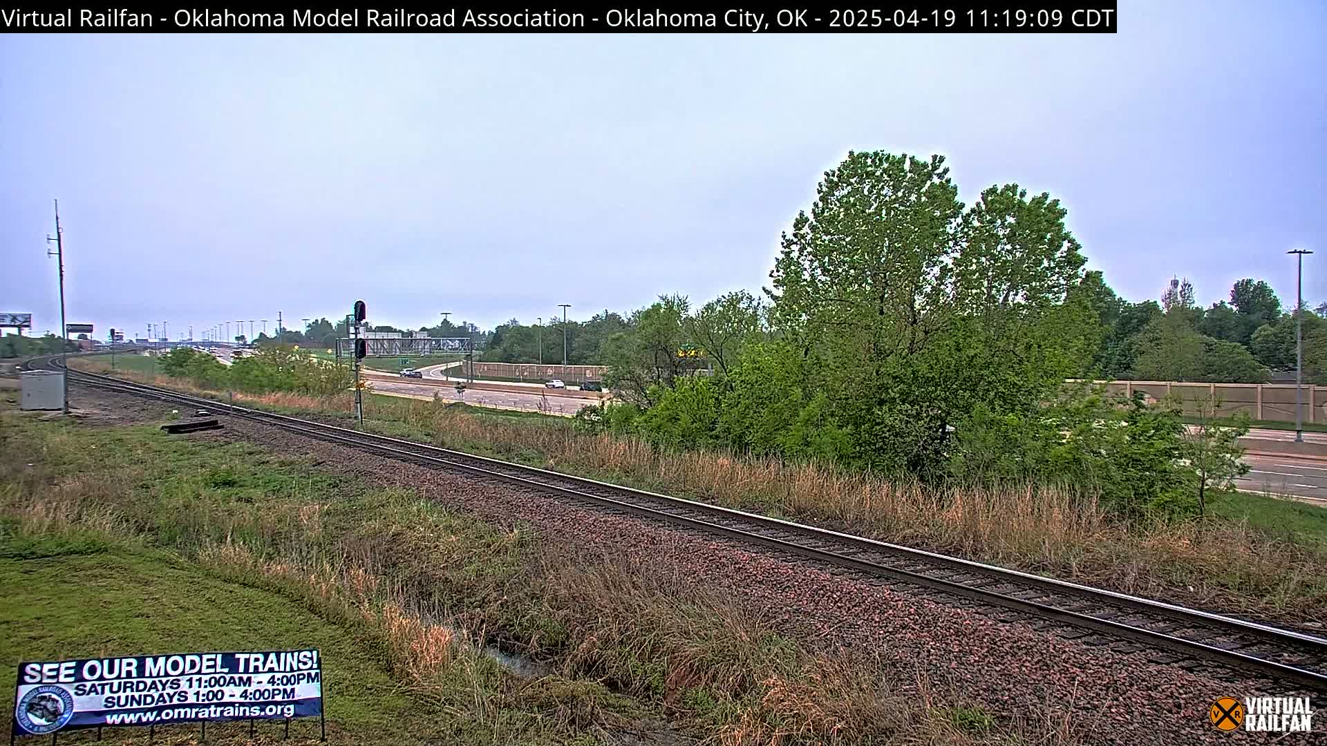 A railroad track runs alongside a highway, with green trees and grass visible under an overcast sky.