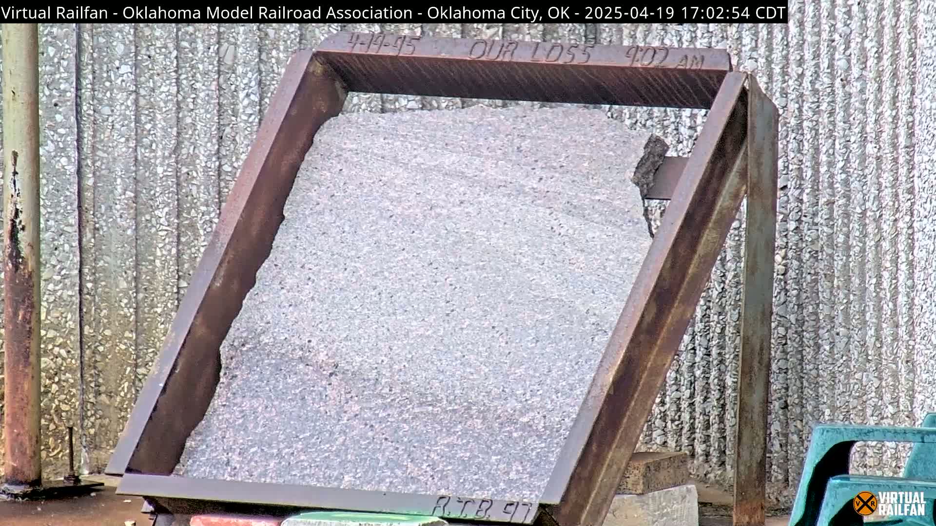 A rusty metal frame holds a pile of light gray gravel against a textured concrete wall outdoors.