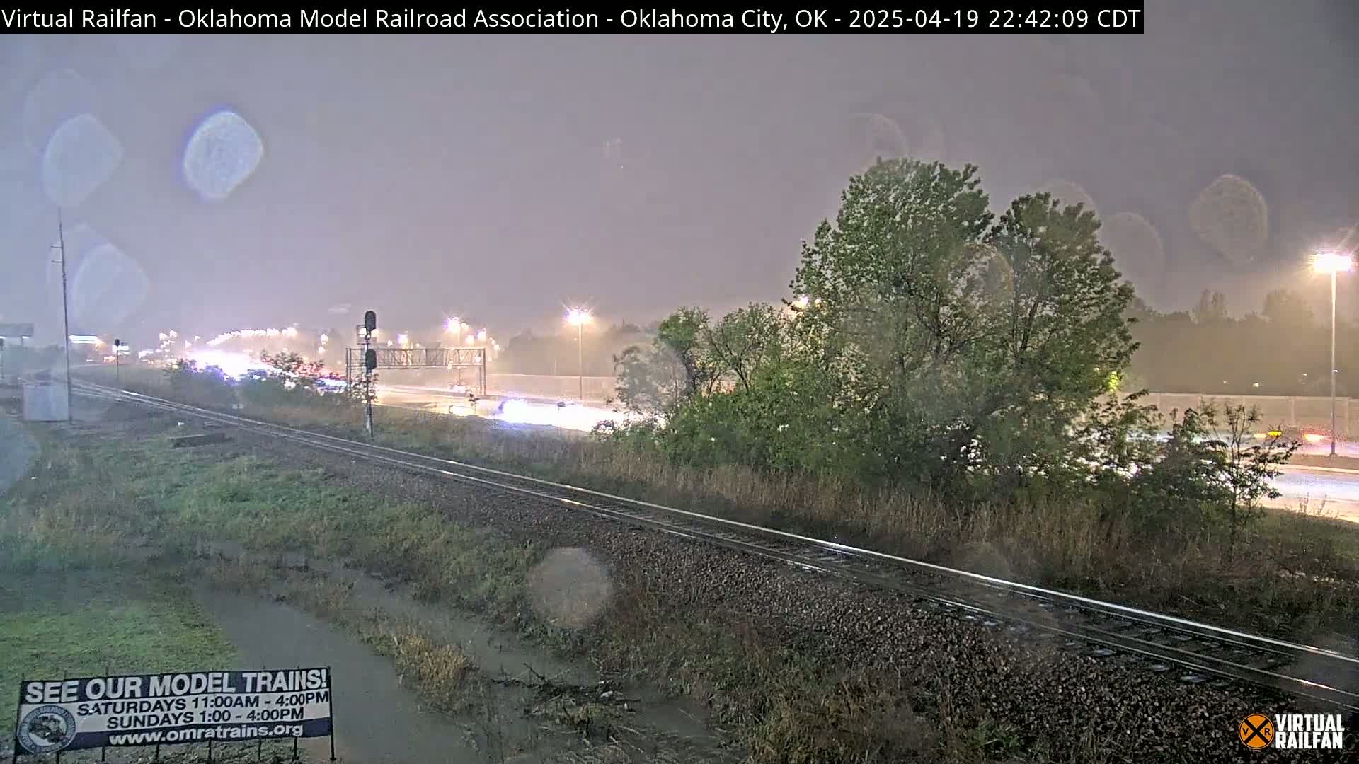 A nighttime view of a rain-slicked highway and railroad tracks, with a few cars visible, and trees alongside.