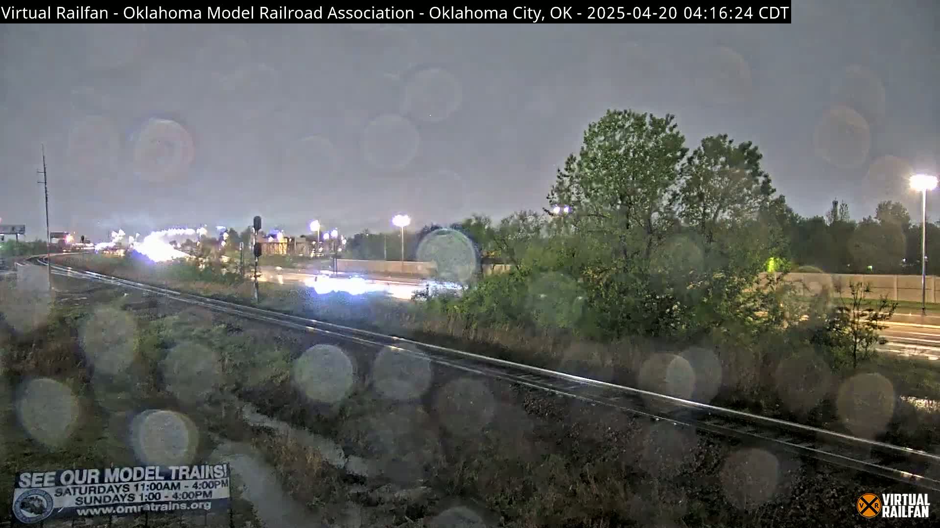 Rain falls on a nighttime scene of railroad tracks next to a roadway with sparse traffic and vegetation.