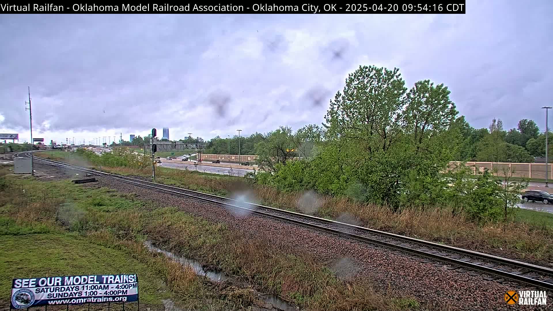 A view of railroad tracks next to a highway on an overcast, rainy day, with green vegetation and trees bordering both sides.