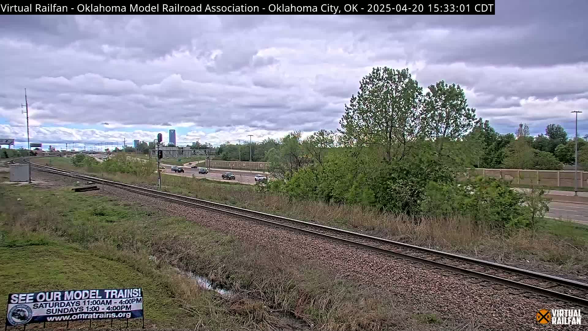 A mostly cloudy sky overlooks a railway track beside a road with sparse traffic, next to a grassy area and a line of trees.