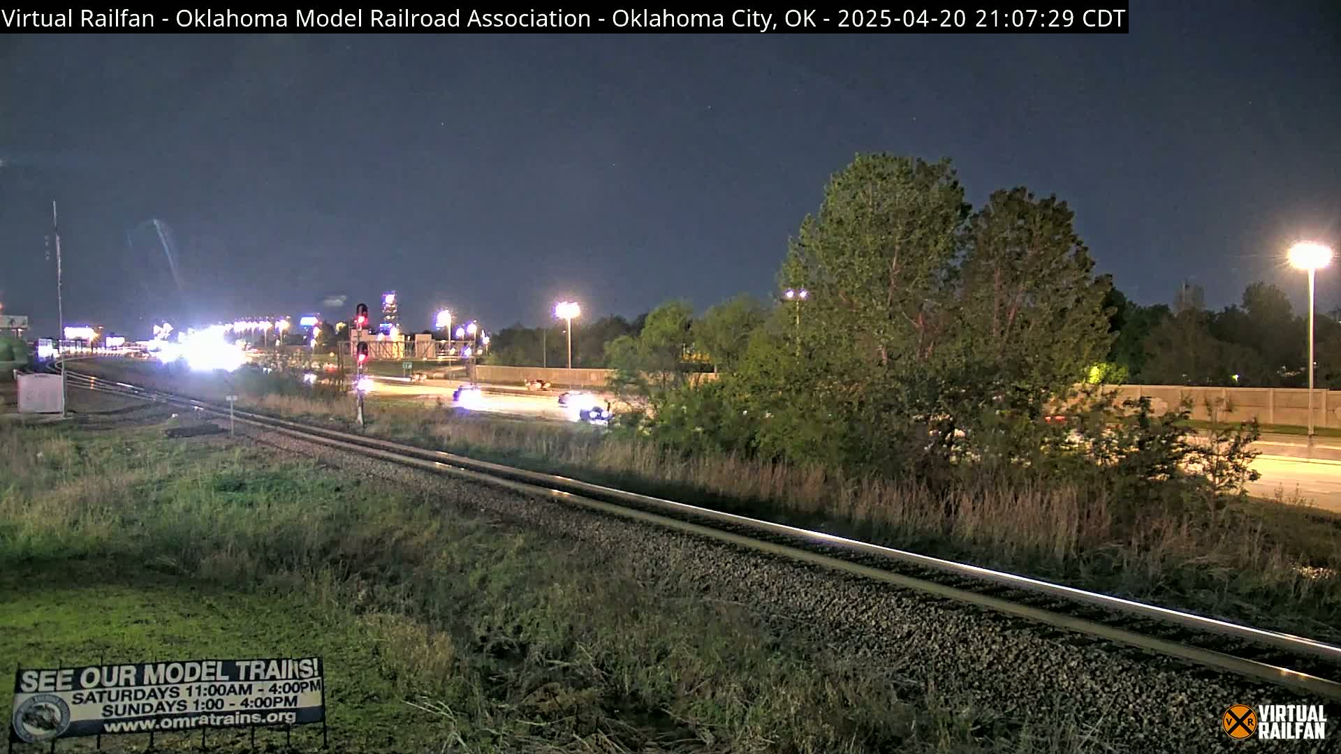 A nighttime view of railroad tracks beside a highway with sparse traffic under a clear sky.