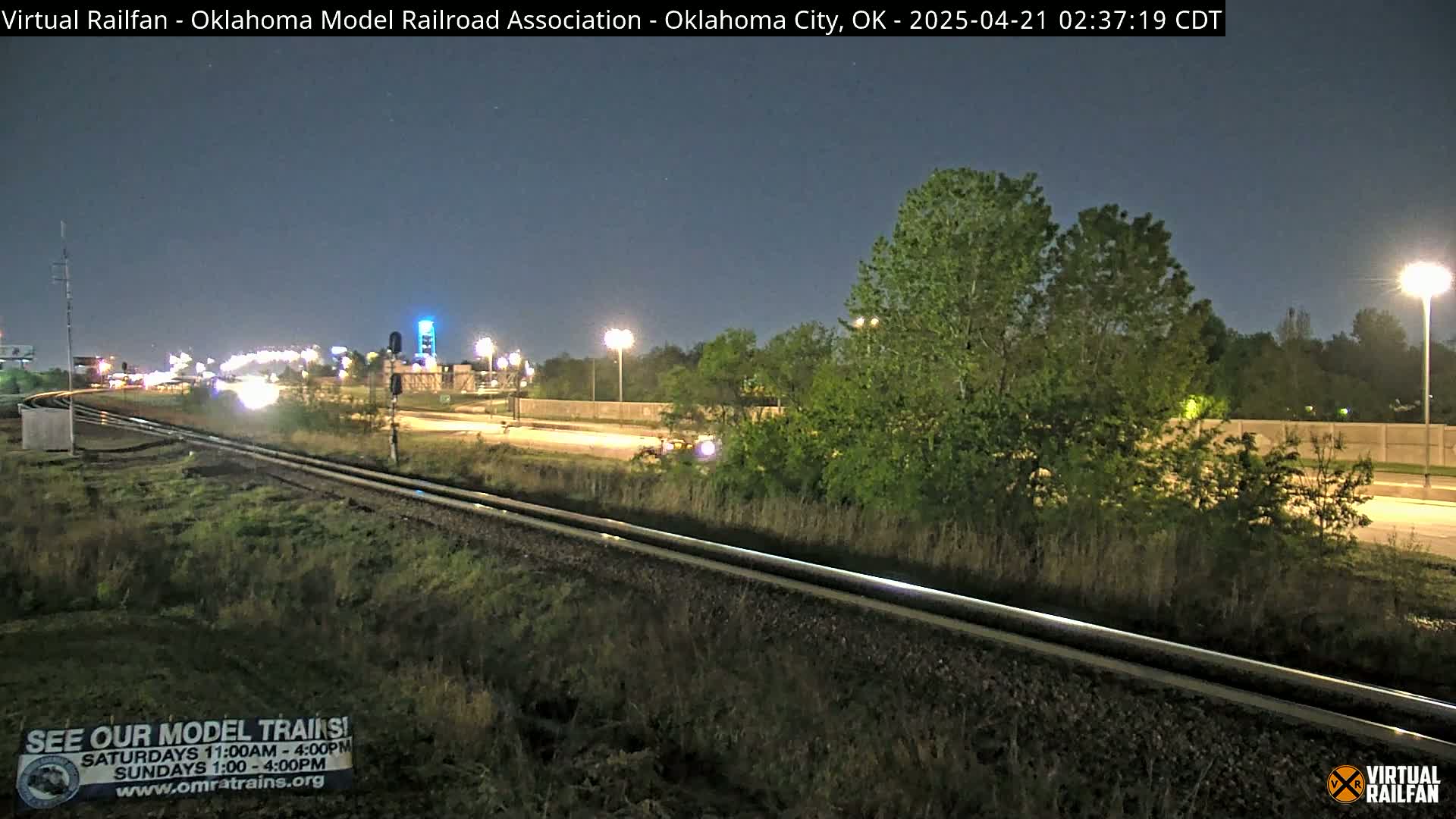 A nighttime view of railroad tracks next to a highway, with trees and streetlights visible under a clear sky.