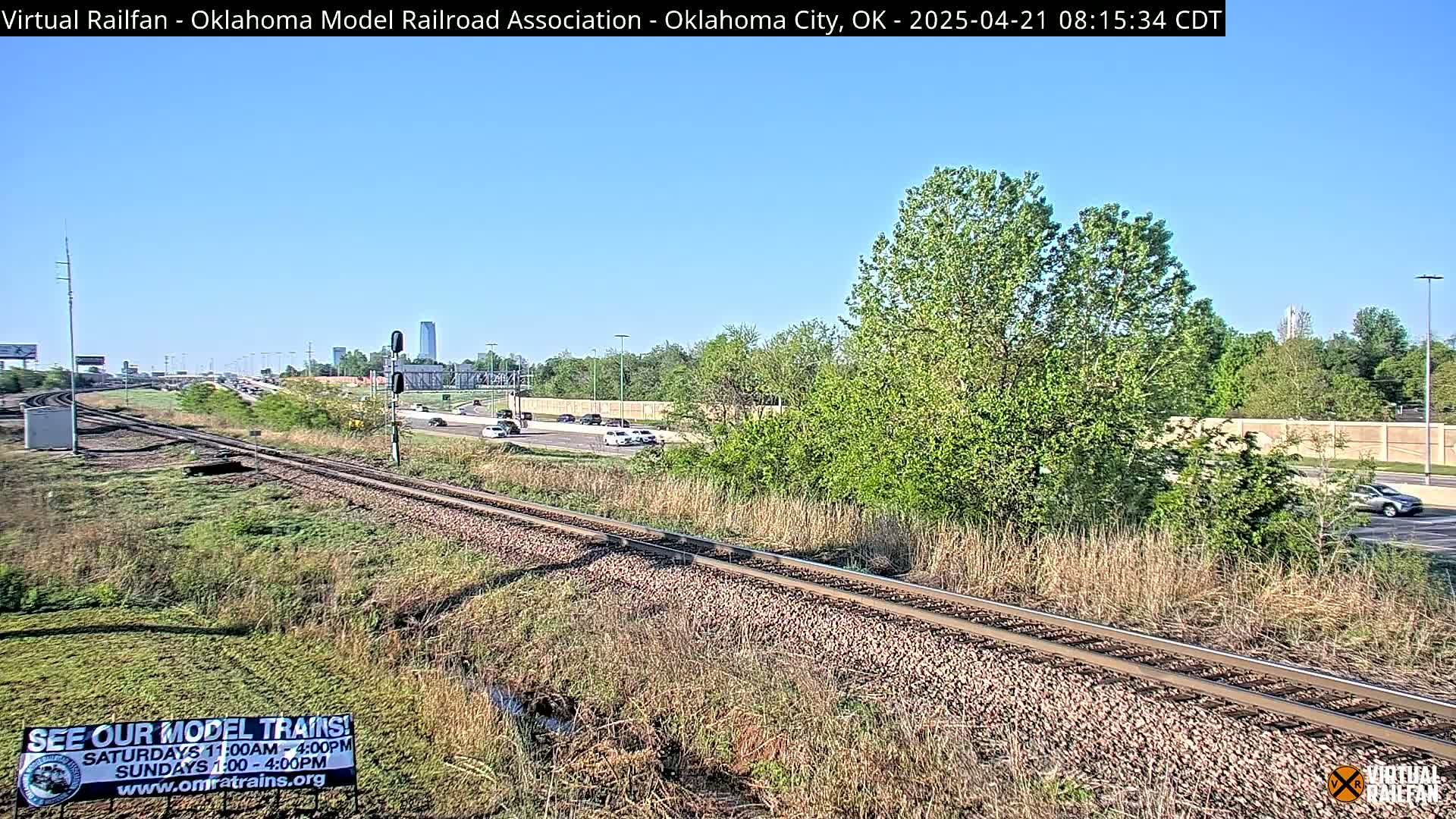 A sunny day shows a railroad track next to a highway with cars driving on it, surrounded by green vegetation and a few tall buildings in the distance.
