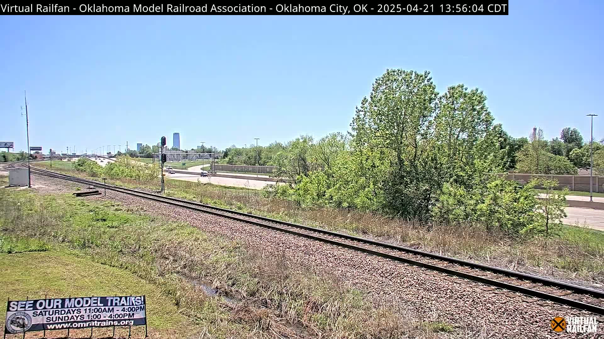 A train track runs alongside a highway, with green vegetation and a clear blue sky overhead.