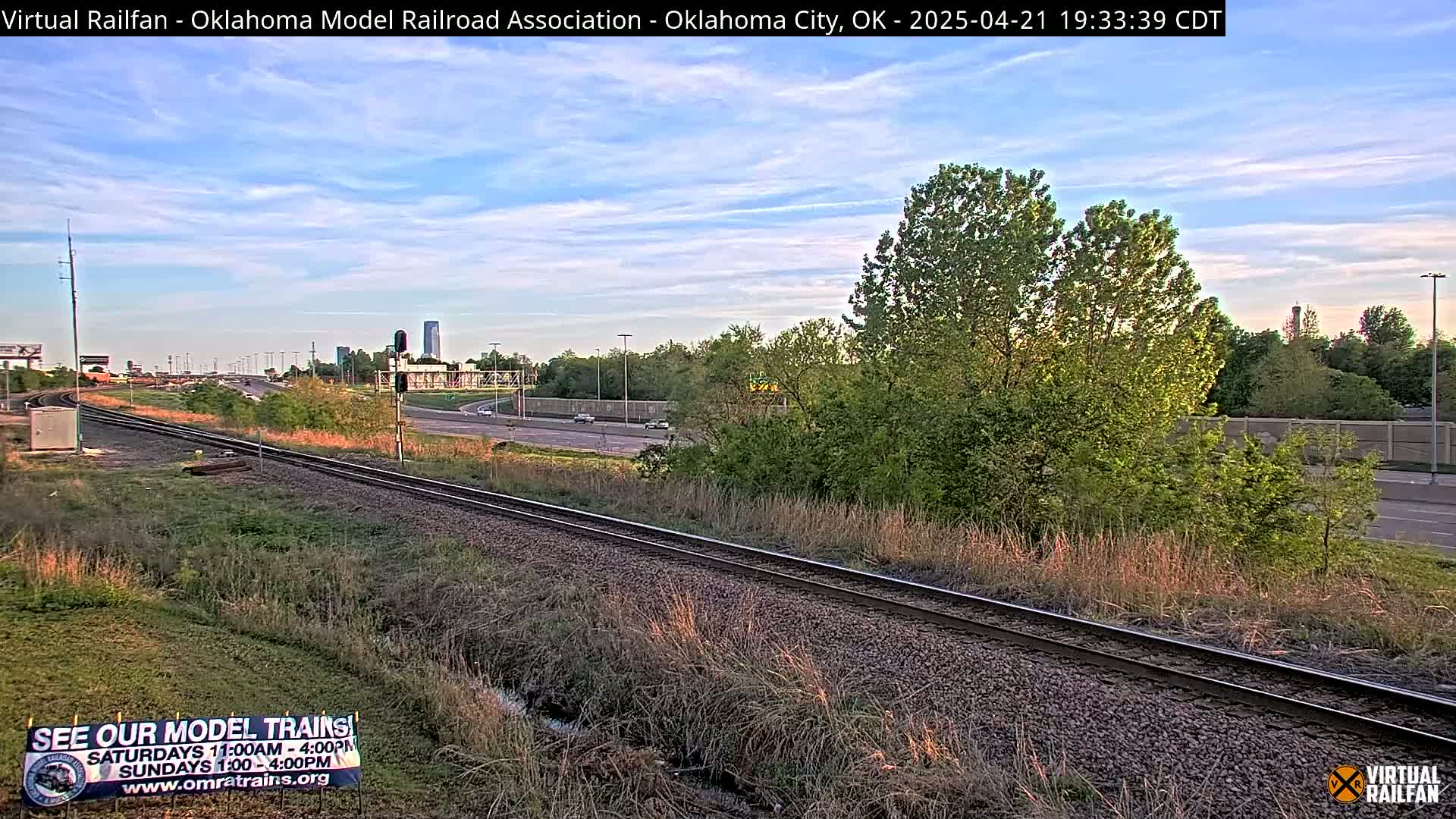 Railroad tracks beside a highway, under a partly cloudy evening sky, with green trees and grass in the foreground.