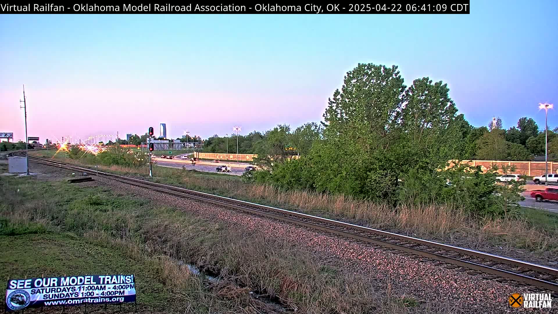 A train track runs parallel to a highway with light traffic under a twilight sky.