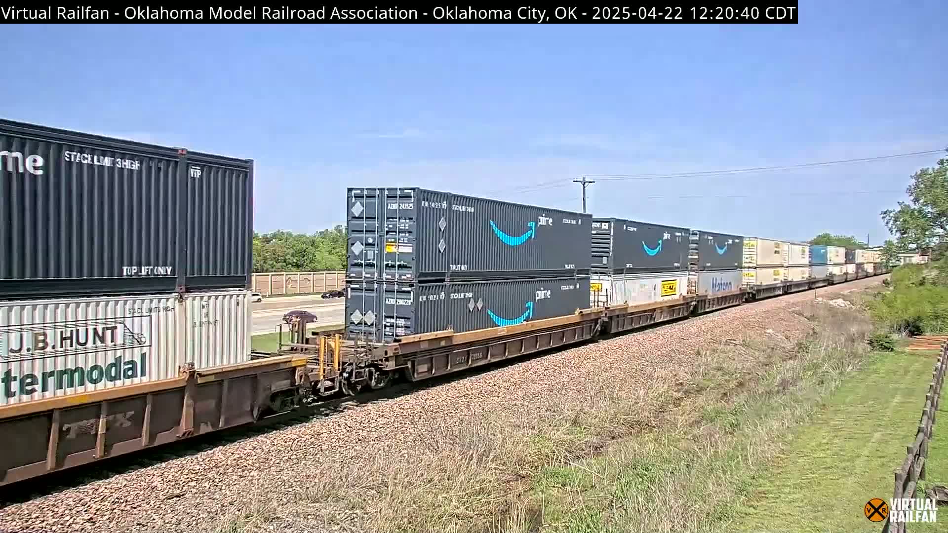 A long freight train carrying numerous shipping containers, some bearing the Amazon Prime logo, travels along a railway track under a clear, sunny sky.