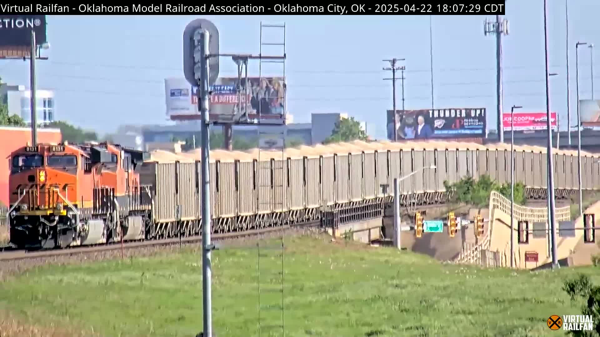 A long freight train with an orange locomotive pulls numerous tan boxcars across a grassy landscape under a clear sky.