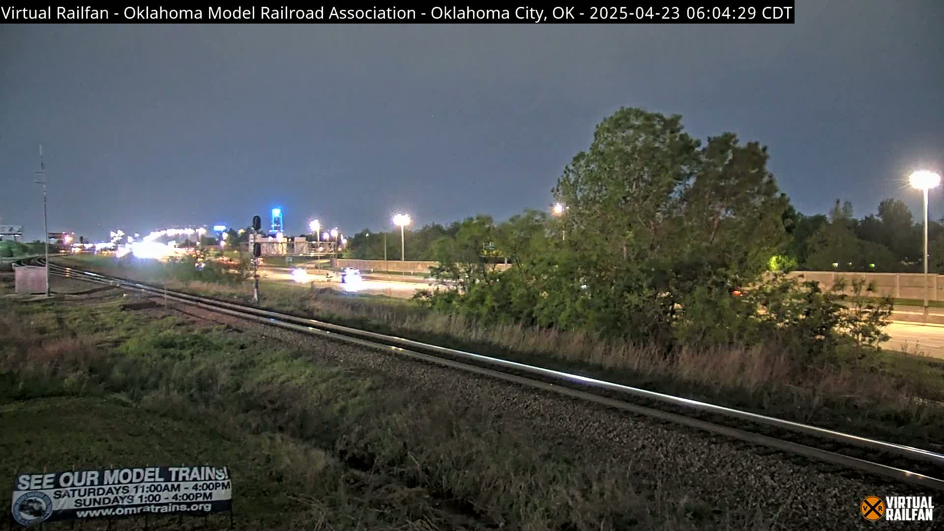 Under a twilight sky, railroad tracks run alongside a highway with sparse nighttime traffic, bordered by vegetation.