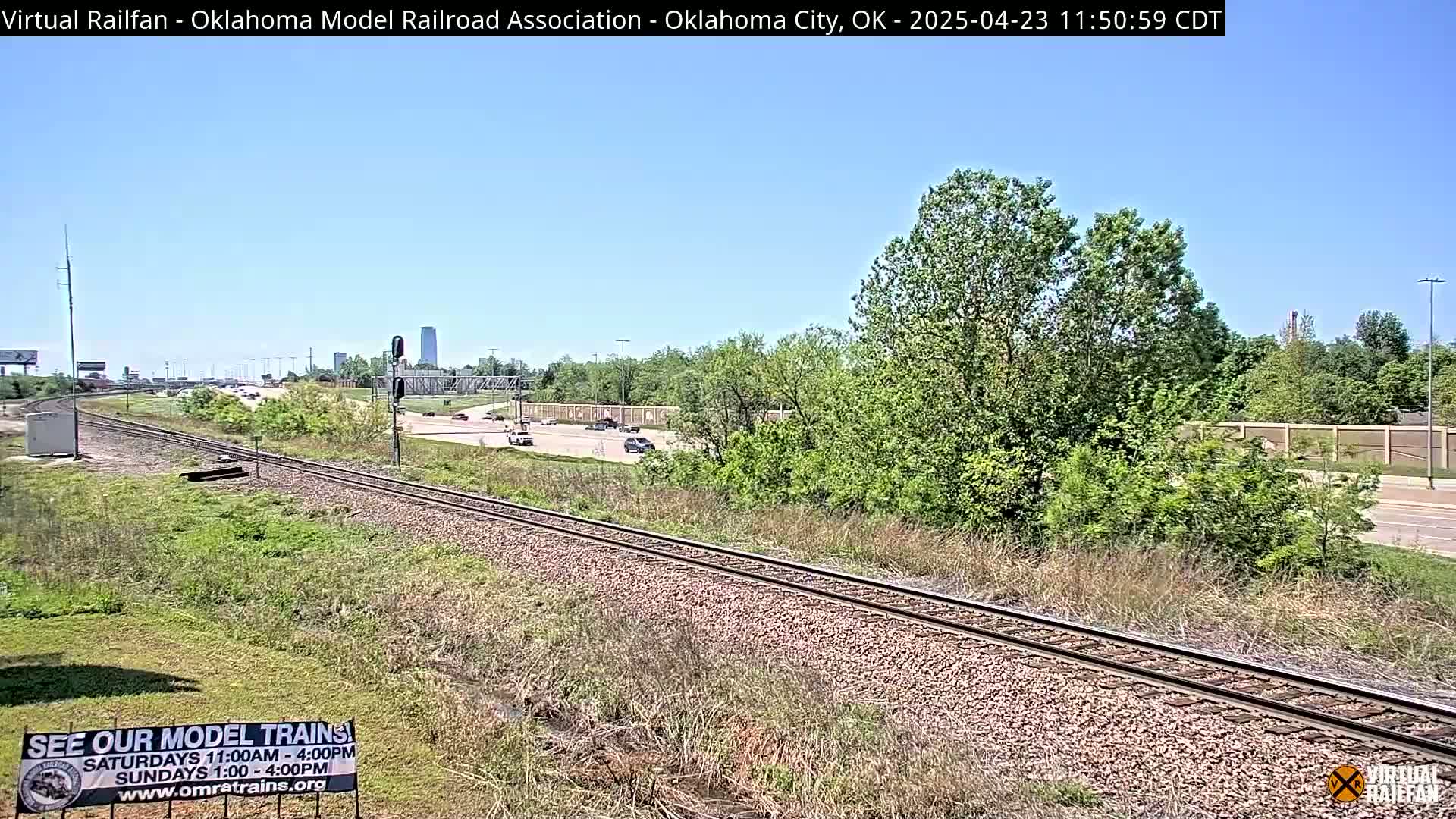 A train track runs alongside a highway, with green vegetation and a few cars visible on the road under a clear blue sky.