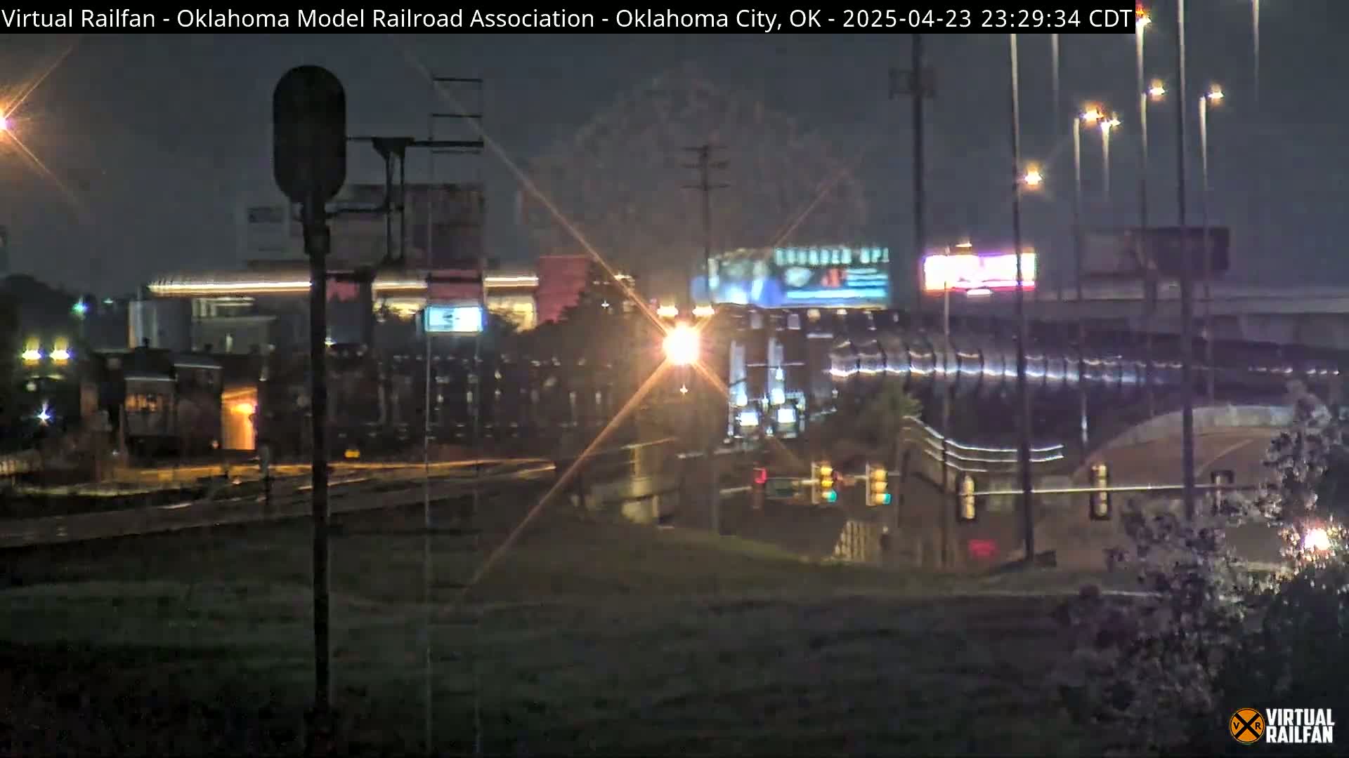 A nighttime view of a train passing by under bright streetlights in the rain.