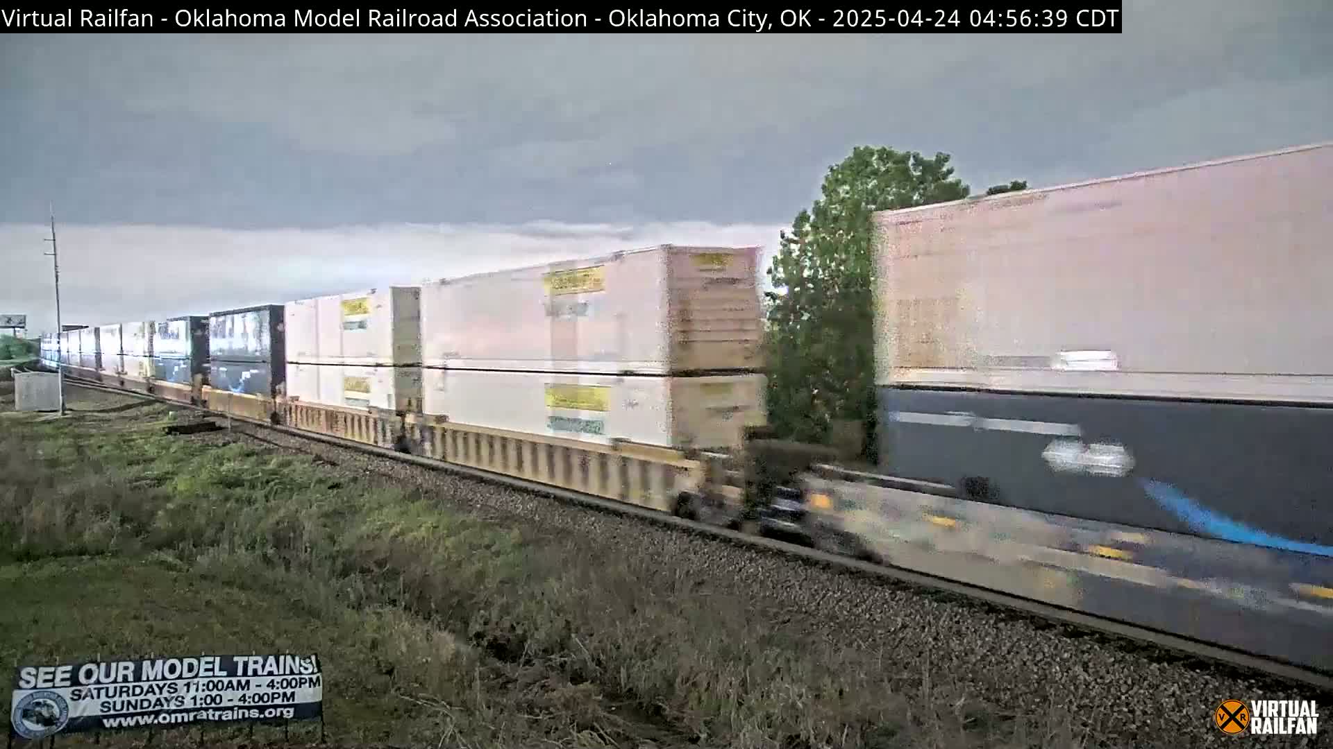 A long freight train with many white and tan shipping containers moves along a railway track under an overcast sky.