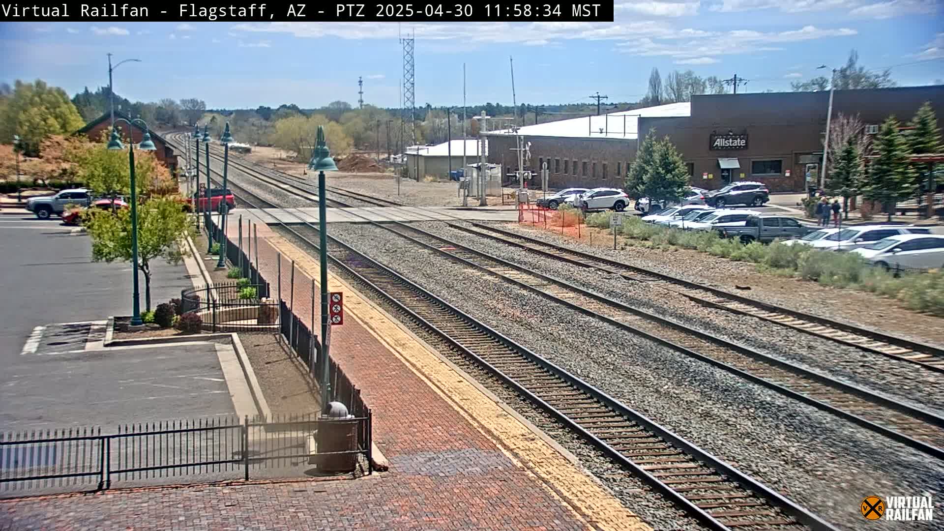 A sunny day reveals a train station with multiple tracks, a parking lot with cars, and a brick pathway leading to a building in the background.