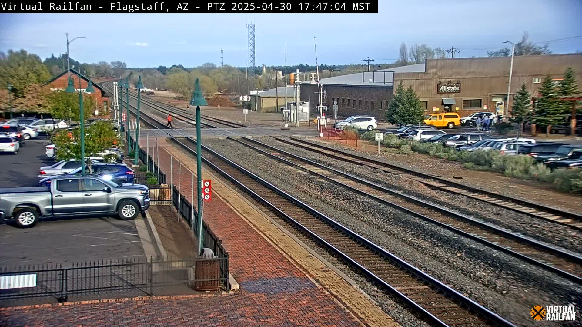 A railroad crossing with multiple train tracks, a parking lot filled with cars, and a few buildings in the background under a mostly clear sky.