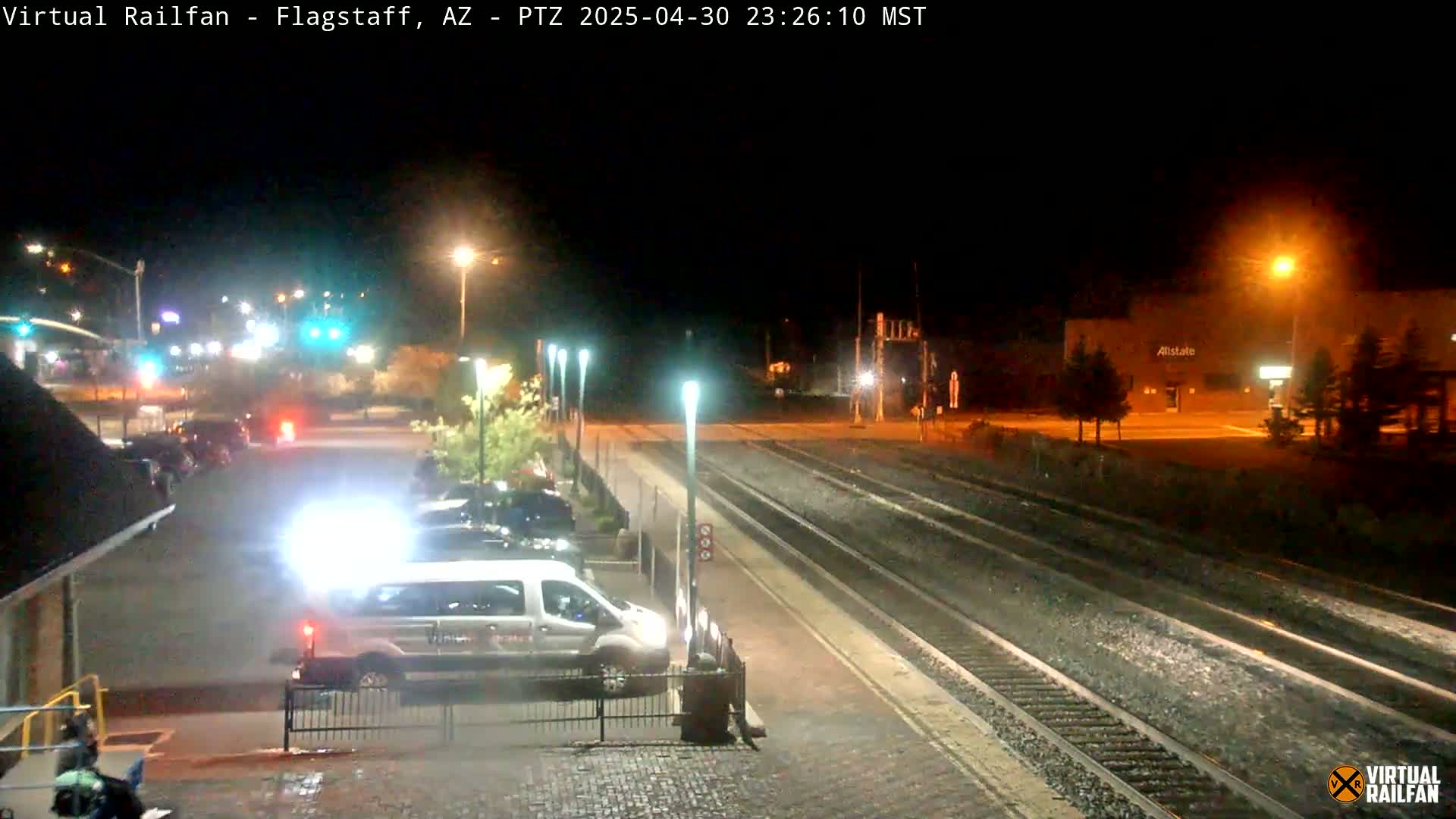 A nighttime view of a train station platform with a van parked nearby, and train tracks extending into the distance under artificial lighting.