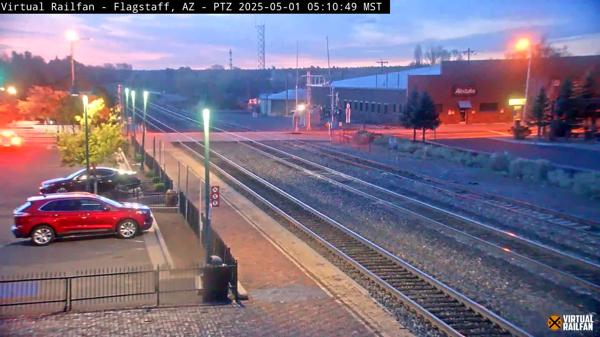 A twilight view of a train station with several train tracks, a red SUV parked nearby, and a few buildings in the background.